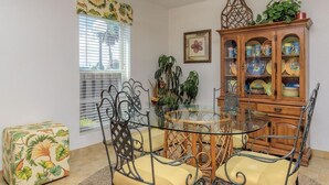 A dining area with a glass-top table, metal chairs, a wooden cabinet with dishes, and a window with blinds. - Unit 101 Villa Del Sol (South Padre Island)