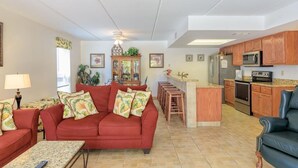 A living room with a red sofa, a coffee table, and a kitchen area with wooden cabinets and a microwave. - Unit 101 Villa Del Sol (South Padre Island)