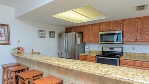 A kitchen with granite countertops, wooden cabinets, stainless steel appliances, and a bar area with stools. - Unit 101 Villa Del Sol (South Padre Island)