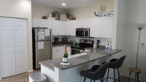 A modern kitchen with white cabinets, a stainless steel refrigerator, a microwave, and a sink. There are two black bar stools and a bottle of champagne on the counter.