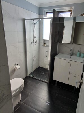 A modern bathroom with a glass-enclosed shower, a toilet, a sink with a mirror, and a window above the sink. - Wallis View - Lake and Sunset Views (Forster)