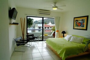 A hotel room with a bed, a TV, a chair, a table, a ceiling fan, and a window with a view of buildings and hills. - Best Value in Heart of Old Town - Modern Architecture -- Free WiFi (Puerto Vallarta)