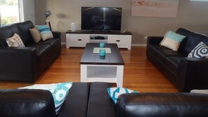 Un salon avec deux canapés noirs, une table basse, un téléviseur posé sur un meuble blanc et un panneau décoratif représentant une plage accroché au mur.