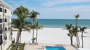 A pool area with a view of the beach and ocean, surrounded by palm trees and a building with balconies.