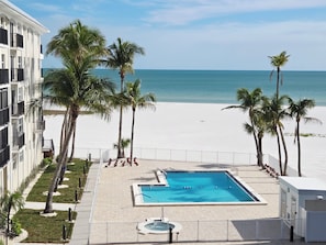 Área da piscina com vista para a praia e oceano, cercada por palmeiras e um edifício com varandas.
