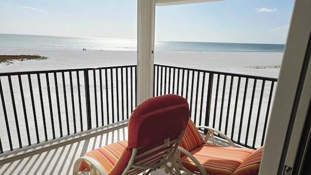 A balcony with a view of the beach, featuring two striped chairs and a white metal railing.