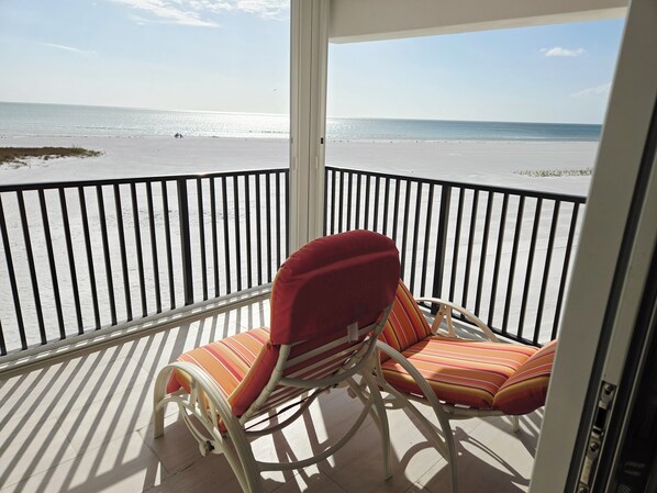 Uma varanda com vista para a praia, com duas cadeiras listradas e um corrimão de metal branco.