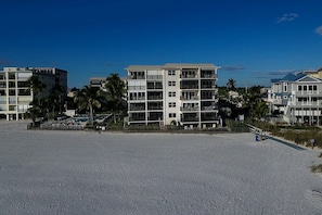 Um hotel na praia com várias varandas, uma praia arenosa em primeiro plano e palmeiras ao fundo.