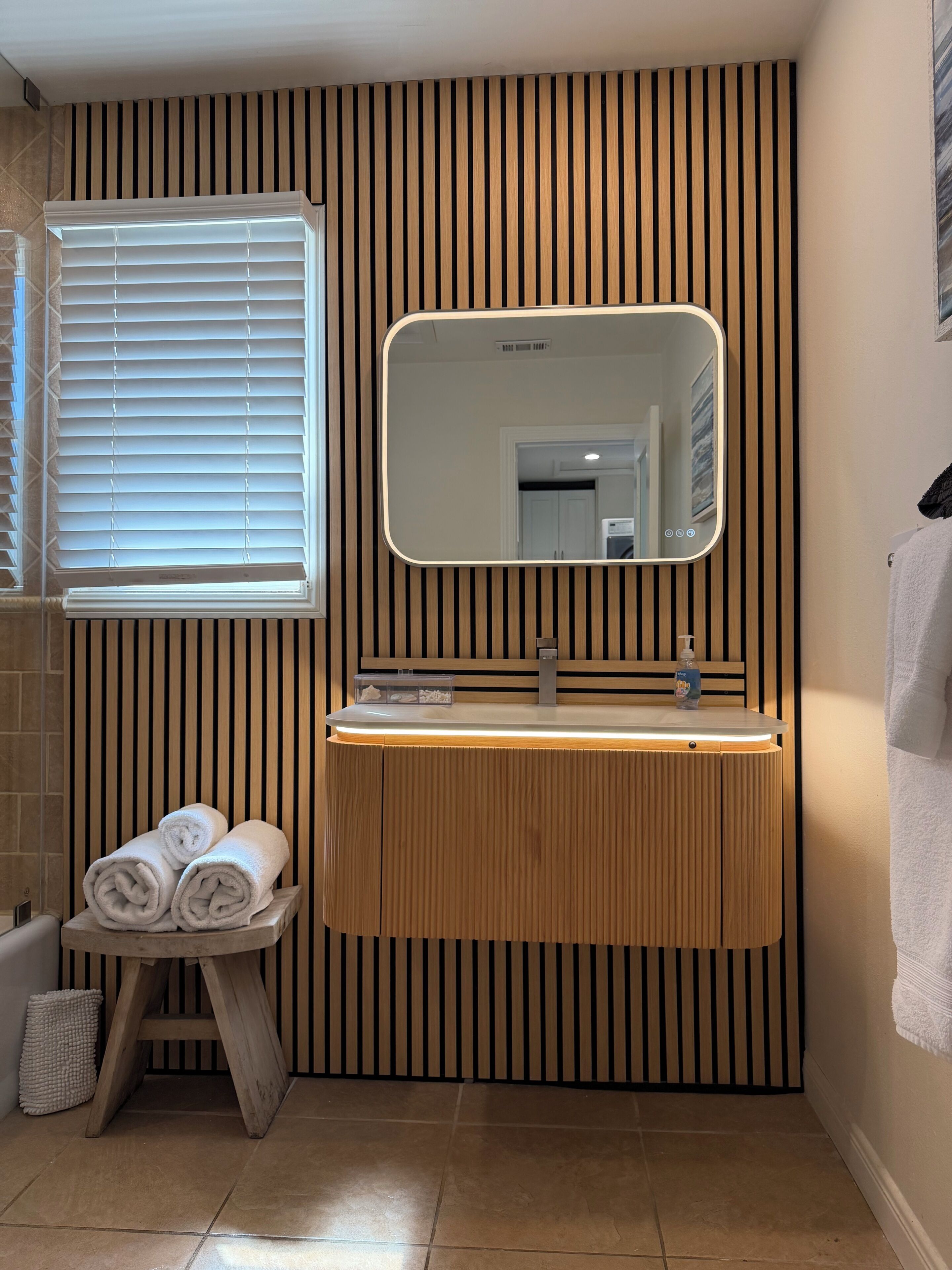 Un baño con pared de paneles de madera, una ventana con persianas, un espejo, un lavamanos con dispensador de jabón y toallas enrolladas sobre un taburete de madera.