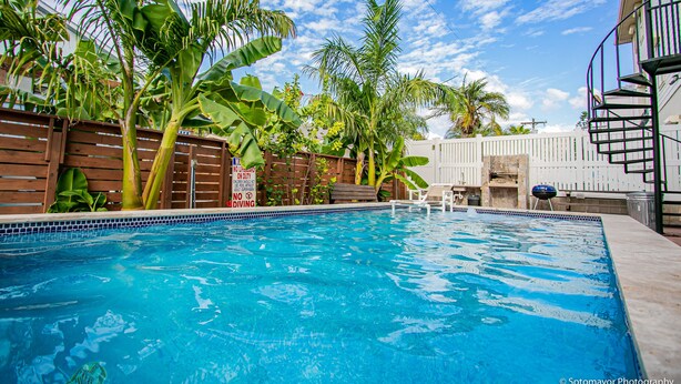 Une piscine entourée d’une clôture en bois, avec des palmiers et un bâtiment blanc en arrière-plan.