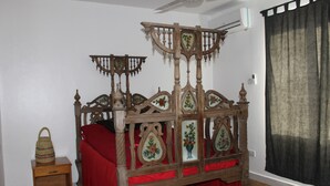 A wooden four-poster bed with intricate carvings and floral designs, a small wooden nightstand with a woven basket, and a red bedspread. - Homely & Friendly property (DSM)
