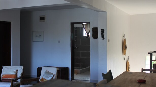 A spacious room with a large wooden dining table, a sofa, and a chair. There is a doorway leading to a bathroom with a tiled shower area. - Homely & Friendly property (DSM)