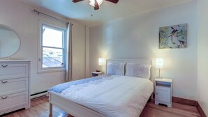 A bedroom with a bed, two bedside tables with lamps, a window with curtains, a ceiling fan, and a wall-mounted artwork.