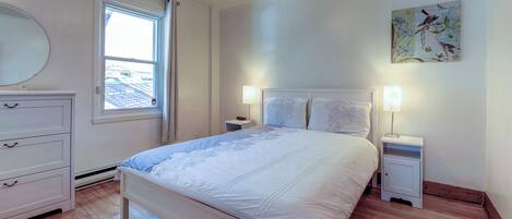 A bedroom with a bed, two bedside tables with lamps, a window with curtains, a ceiling fan, and a wall-mounted artwork.