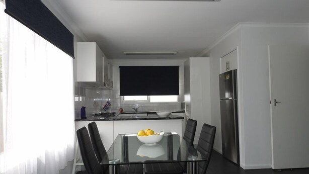 A modern kitchen with a glass dining table, black chairs, and a bowl of fruit.