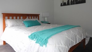 A neatly made bed with a white duvet and teal pillows, a teal throw blanket, and a cityscape painting on the wall.