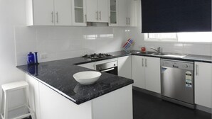 A modern kitchen with white cabinets, a black countertop, a gas stove, an oven, a dishwasher, and a window with a dark blind.
