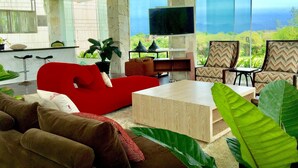Ein modernes Wohnzimmer mit einem roten Sofa, einem Holztisch und zwei gemusterten Sesseln. Ein Flachbildfernseher ist an der Wand befestigt, und große Fenster bieten einen Blick ins Freie.