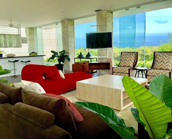 Una sala de estar moderna con un sofá rojo, una mesa de centro de madera y dos sillones estampados. Un televisor de pantalla plana está montado en la pared, y hay ventanas amplias que ofrecen vistas al exterior.