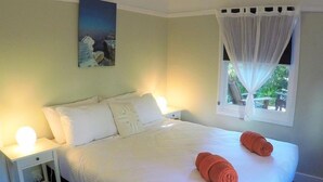 A neatly made bed with white linens and two rolled towels, a bedside table with a lamp, a painting on the wall, and a window with a curtain.