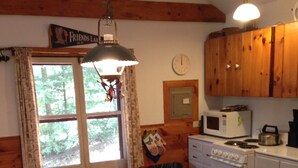 A cozy cabin-style kitchen with wooden cabinets, a dining table with chairs, a hanging light fixture, and a window with curtains. - Beautiful lakeview cottage on Friends Lake (Chestertown)