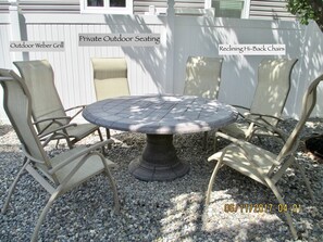 Un barbecue Weber rond, installé à l’extérieur, est entouré de fauteuils inclinables à haut dossier. L’ensemble est posé sur du gravier, avec une clôture blanche en arrière-plan.