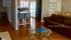 A living room with a queen sleeper sofa, a glass coffee table, and a dining area with a kitchen in the background.
