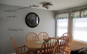 Un coin repas comprenant une horloge, des chaises en bois et une table. On y trouve également un luminaire au plafond et une fenêtre avec des stores.