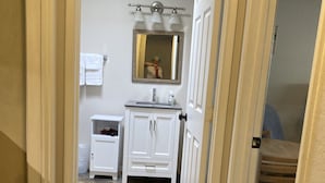Un baño con tocador blanco, un espejo y un lavabo. Hay un perchero en la pared y un cubo de basura. El piso tiene azulejos con diseño.