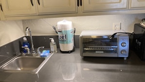 Una cocina con un lavamanos de acero inoxidable, un horno tostador Black+Decker, un paquete grande de toallas de papel, una botella de detergente para platos Dawn y una botella de jabón líquido para manos.