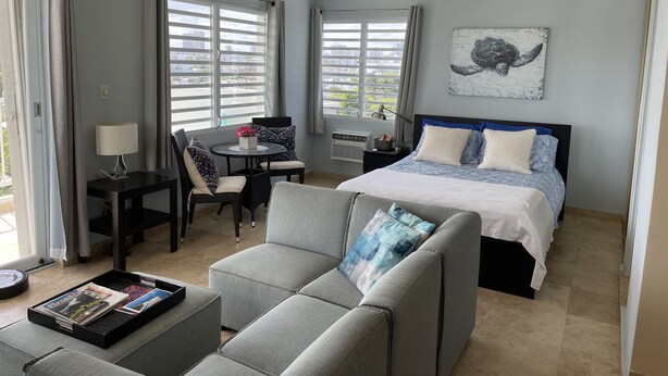 Una habitación de hotel moderna con cama, sofá, escritorio con lámpara y vistas a la ciudad a través de amplios ventanales.