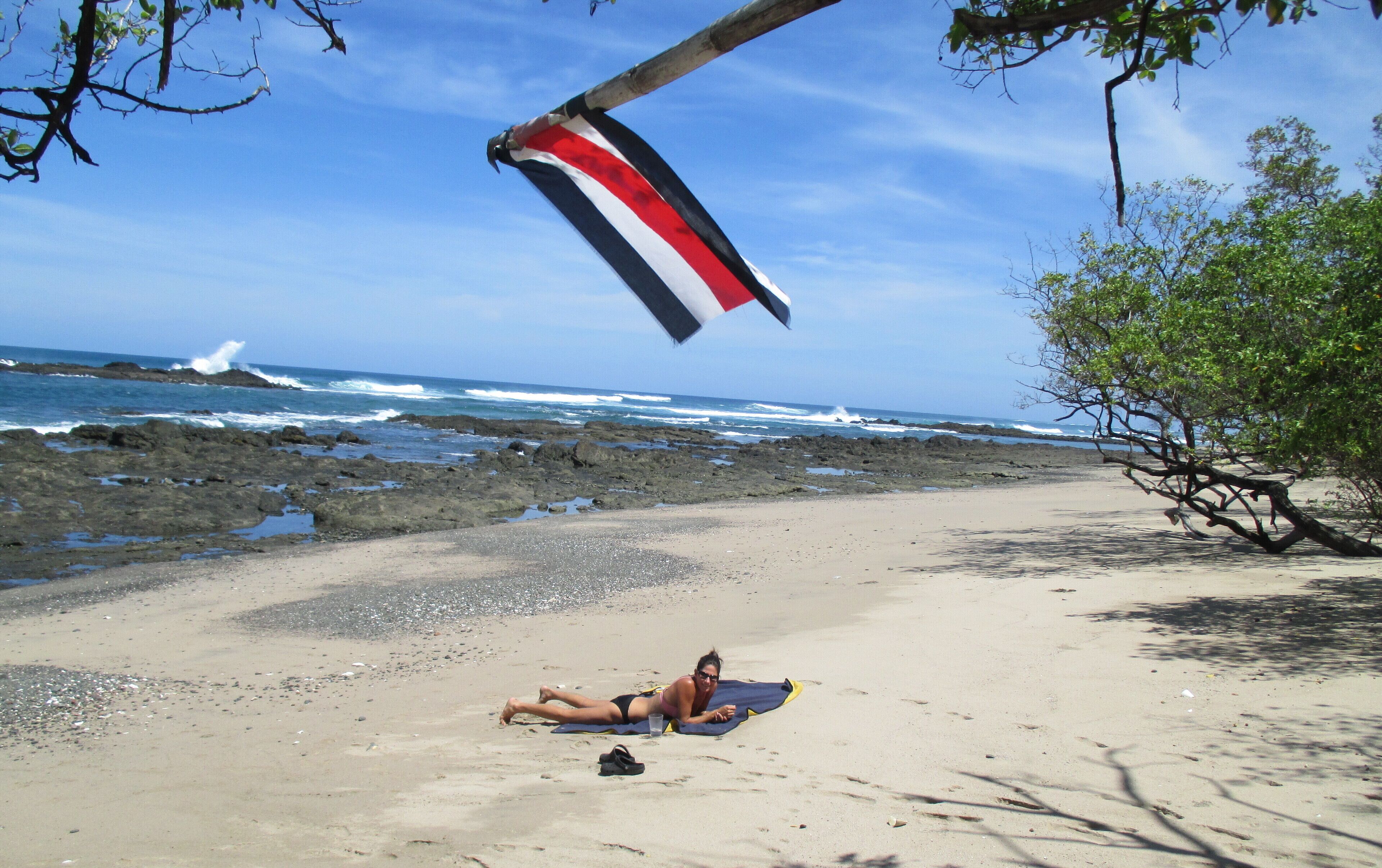 A beach with a person lying on a towel, a flag, and rocky shoreline.