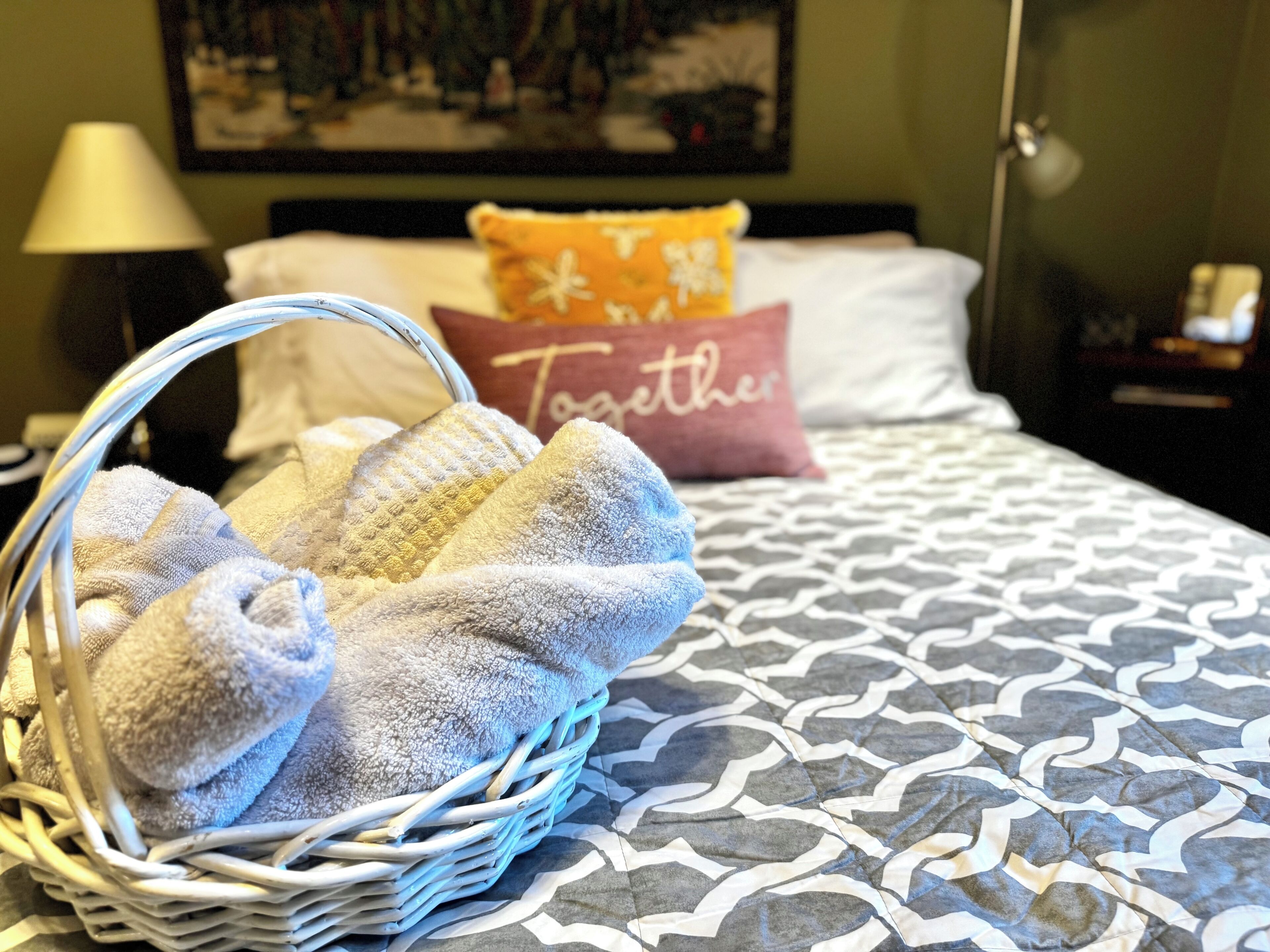 A neatly made bed with patterned bedding, a basket of folded towels, and a pillow with the word "Together".