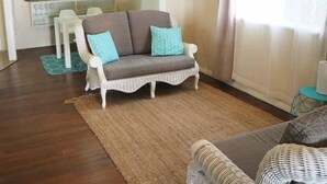 A living room with a white wicker sofa, a brown sofa with blue cushions, a wooden floor, and a dining area in the background. - Feather Reef Cottage (Flying Fish Point)