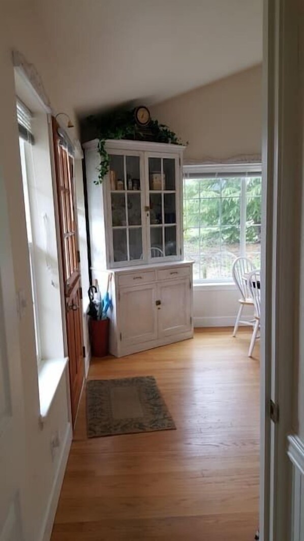 A room with a wooden floor, a white cabinet with glass doors, a chair, and a window with a view of greenery. - Charming, Private Cottage near I-5 and Golf Course. (Myrtle Creek)
