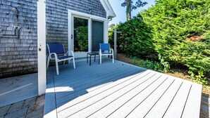 A wooden deck with two chairs and a table, a sliding glass door, and a well-maintained garden with a large evergreen tree.
