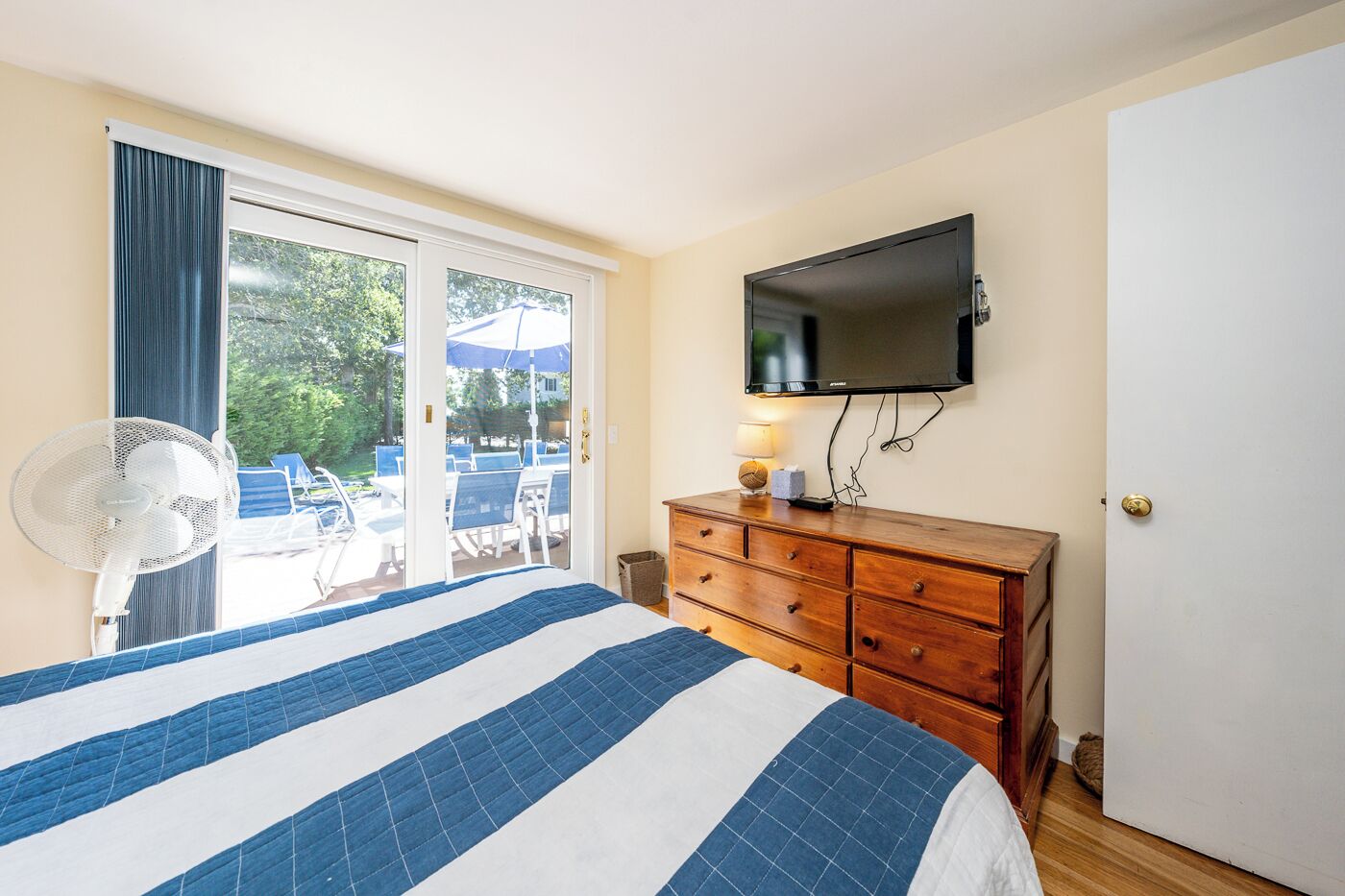 Een hotelkamer met een bed, een houten dressoir, een televisie, een ventilator en een schuifdeur naar een buitenruimte met stoelen en parasols.