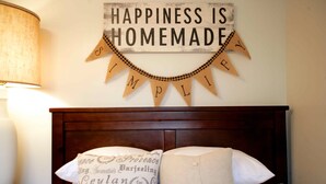Une chambre avec une tête de lit en bois, un lit recouvert de linge de lit blanc et une décoration murale sur laquelle on peut lire : « Le bonheur, ça se crée. »