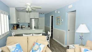 Un salon avec un canapé beige, deux coussins décoratifs, un coin repas avec une table et des chaises, une cuisine équipée d’un réfrigérateur et de placards, et un ventilateur de plafond.