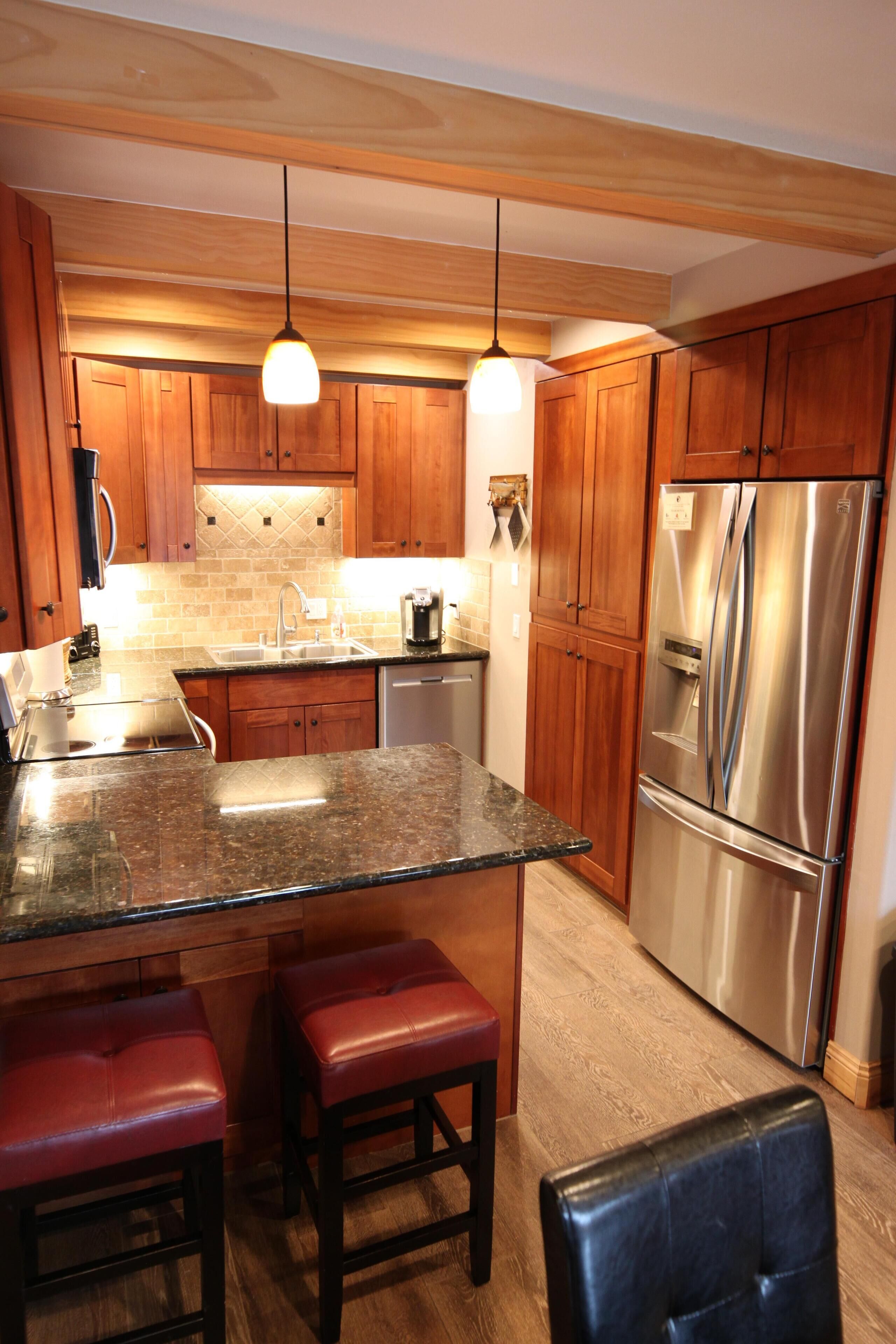 A modern kitchen with wooden cabinets, a stainless steel refrigerator, granite countertops, and pendant lighting.