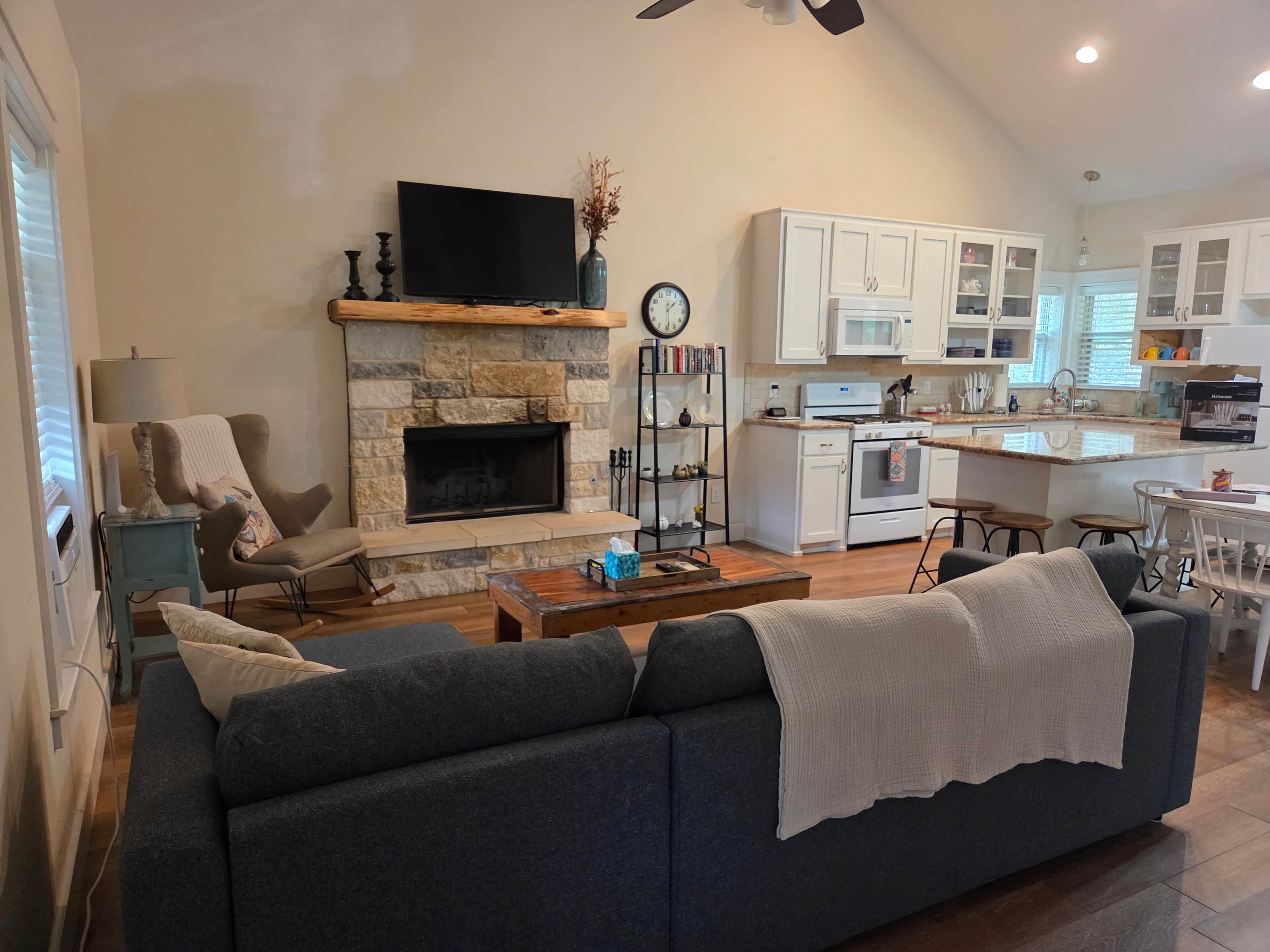 A modern living room with a stone fireplace, a flat-screen TV, a sofa, and a dining area with a kitchen in the background.