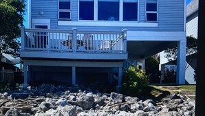 Et hus ved stranden med balkon, to strandstole og en stribet strandmåtte.