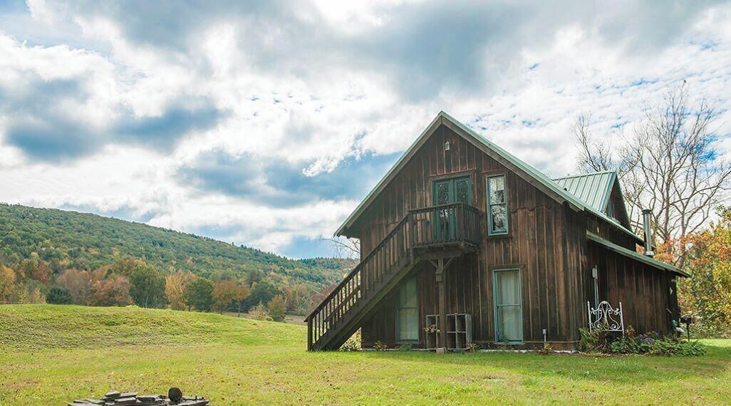 Une grange rustique en bois, dotée d’un balcon, entourée d’un champ verdoyant et avec une colline en arrière-plan.