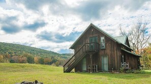 Ein rustikaler Hollbau mit Balkon, umgeben von einer grünen Wiese und einem Hügel im Hintergrund.