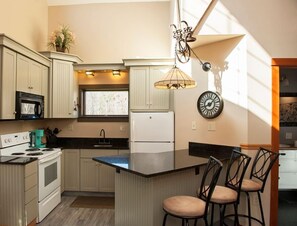 Eine Küche mit weißen Schränken, einem Herd, einem Kühlschrank, einer Spüle und einem Fenster. An der Wand hängt eine Uhr und auf der Arbeitsplatte steht eine Pflanze.