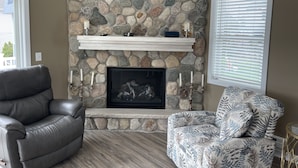 Un salón con chimenea de piedra, un sillón de cuero, un sillón estampado y un estante blanco sobre la chimenea.