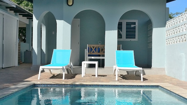 A pool with two sun loungers and a small table, surrounded by a light-colored building with arched doorways. - Villa Valentina Aruba (Noord)