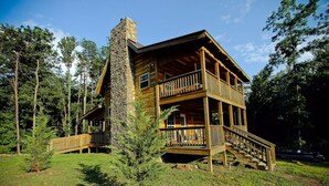 A two-story log cabin with a stone chimney, wooden balconies, and a staircase leading to the upper level.