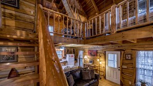A log cabin interior with a loft bedroom, a dining area, and a living room with a leather couch.