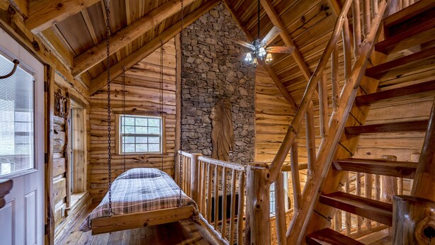 A log cabin bedroom with a loft bed, stone wall, wooden staircase, and a window.